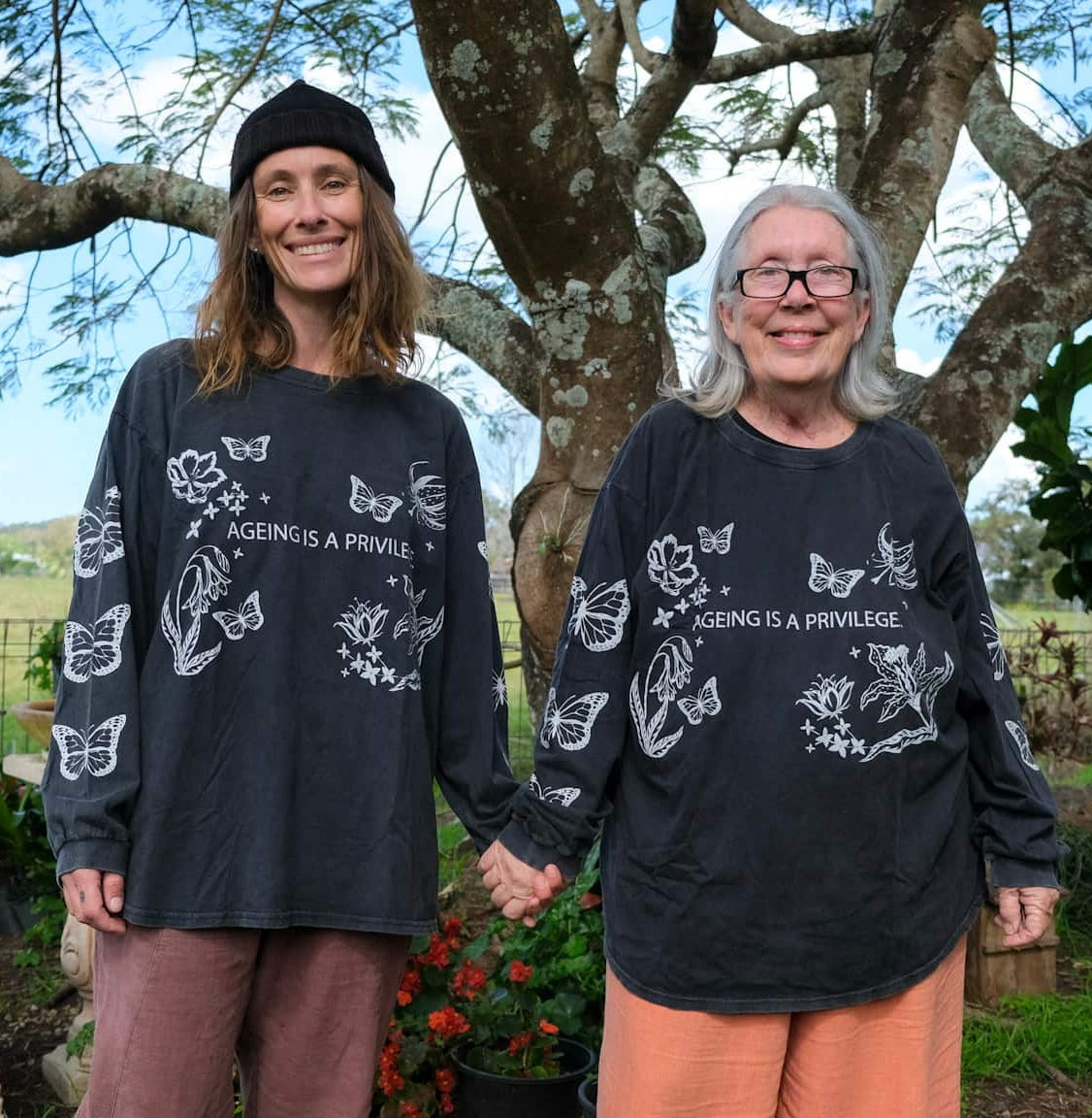 Two women standing outdoors under a large tree, wearing matching black long-sleeve graphic shirts with white butterfly illustrations and the text &