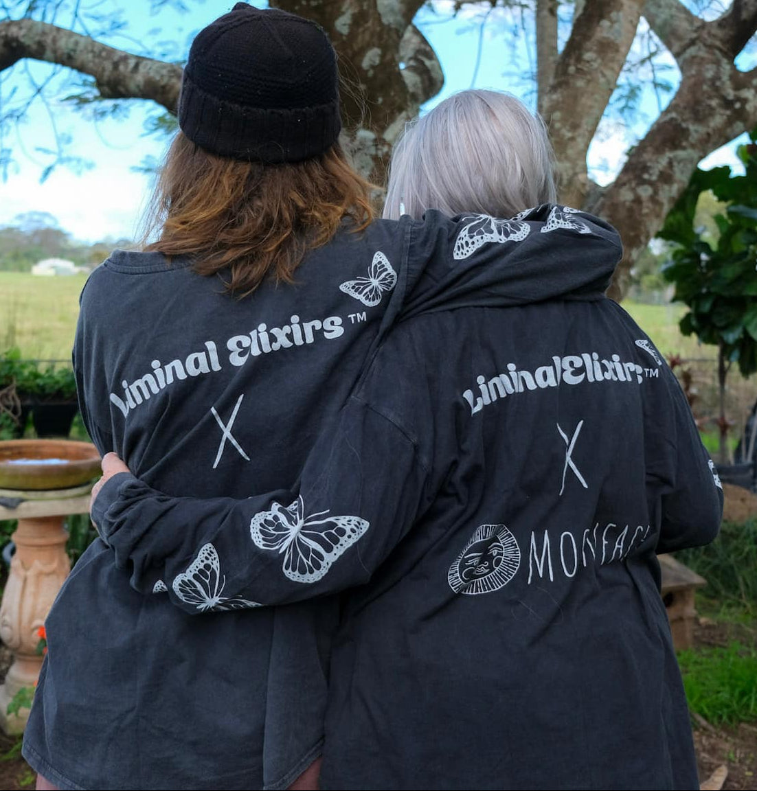 Two people standing outdoors with their backs to the camera, wearing matching black shirts with white graphic text and butterfly designs on the back.