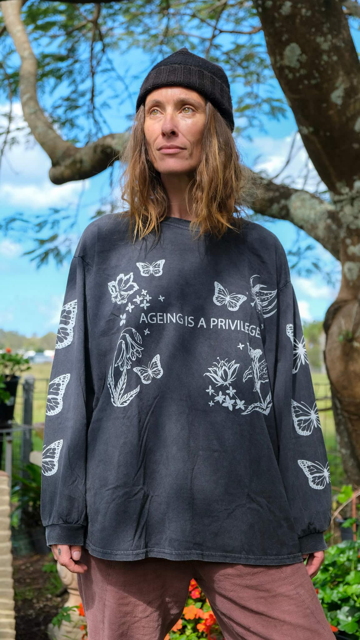 A woman wearing a faded black graphic long-sleeve t-shirt with white butterfly and floral designs, standing outdoors in front of a tree.