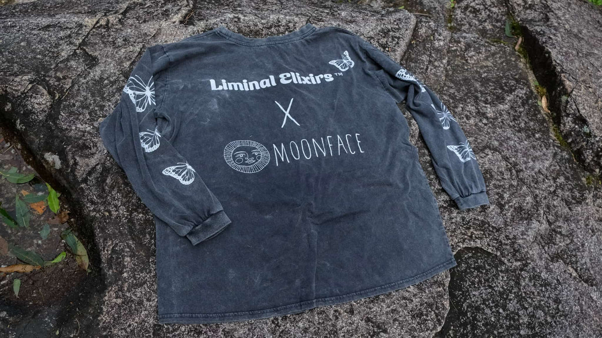 A dark grey washed long-sleeve shirt laid flat on a textured stone surface, featuring white printed graphics including &