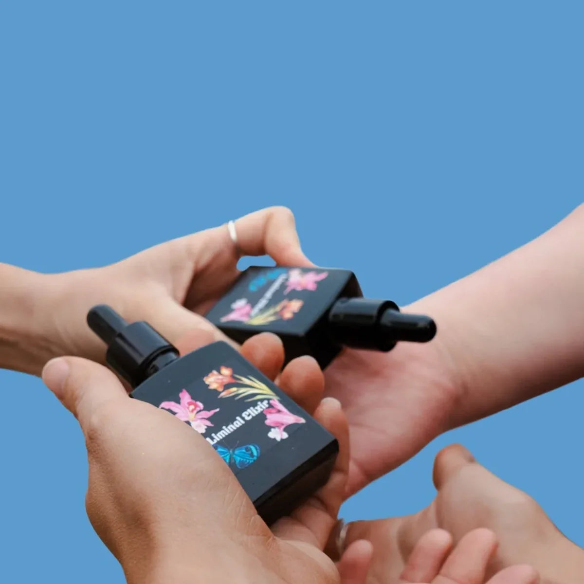 Two black skincare bottles with floral designs held by hands against a blue background