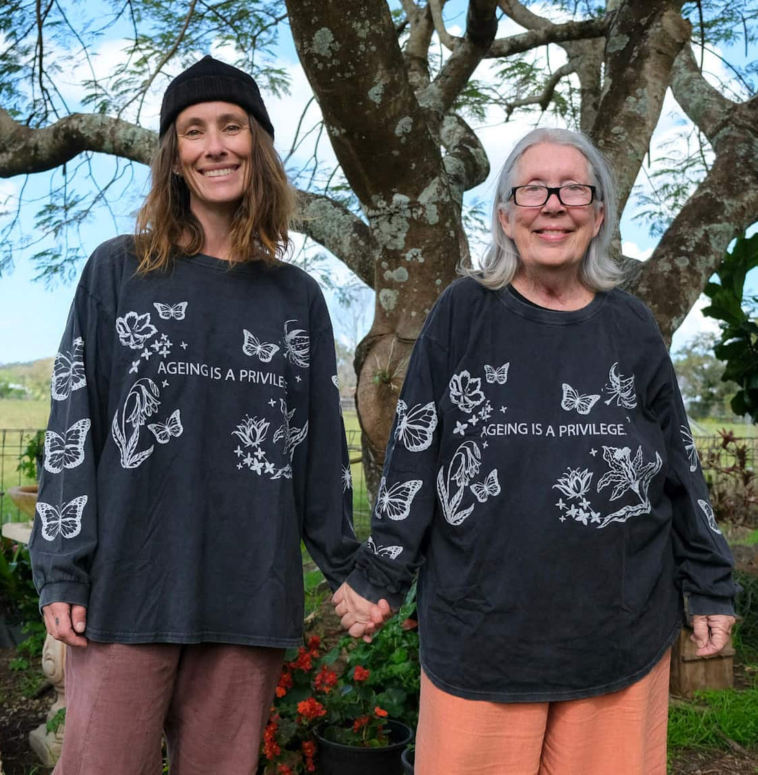 Two women standing outdoors under a large tree, wearing matching black long-sleeve graphic shirts with white butterfly illustrations and the text &