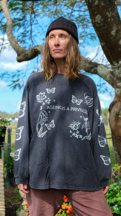 A woman wearing a faded black graphic long-sleeve t-shirt with white butterfly and floral designs, standing outdoors in front of a tree.