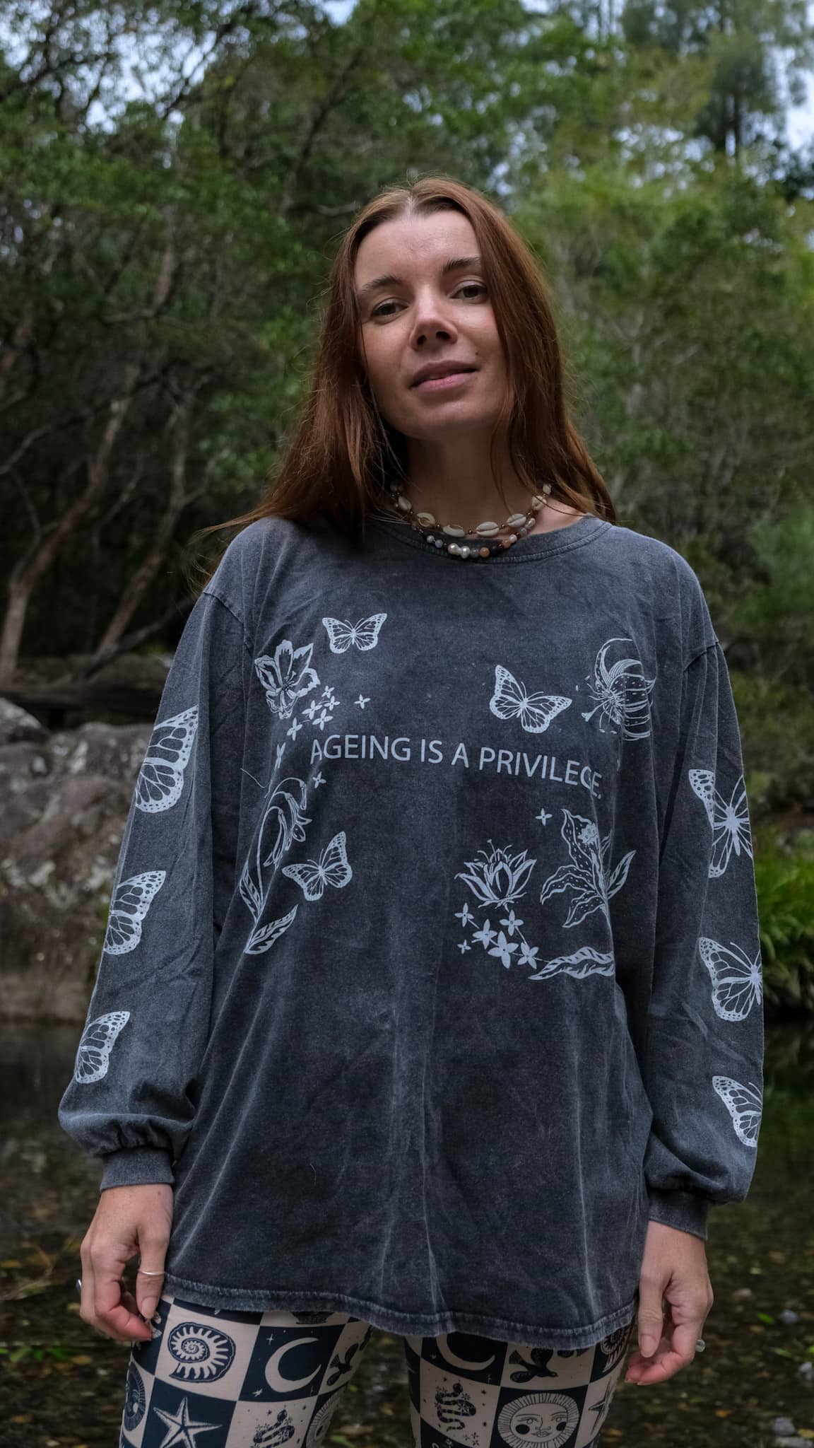 A woman standing outdoors in front of trees and water, wearing a dark charcoal gray long-sleeve t-shirt with white butterfly illustrations and the text &