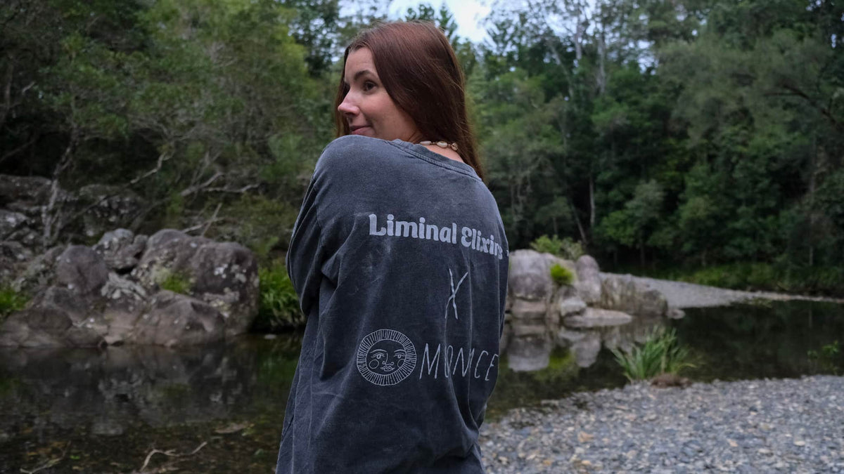A woman with long brown hair stands outdoors by a rocky pond, wearing a dark grey oversized T-shirt with white text reading &