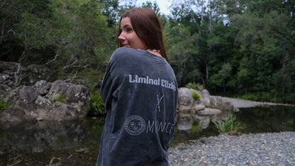 A woman with long brown hair stands outdoors by a rocky pond, wearing a dark grey oversized T-shirt with white text reading &