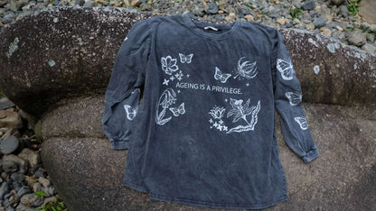 A dark washed charcoal long-sleeve t-shirt with white butterfly and floral illustrations and the text &