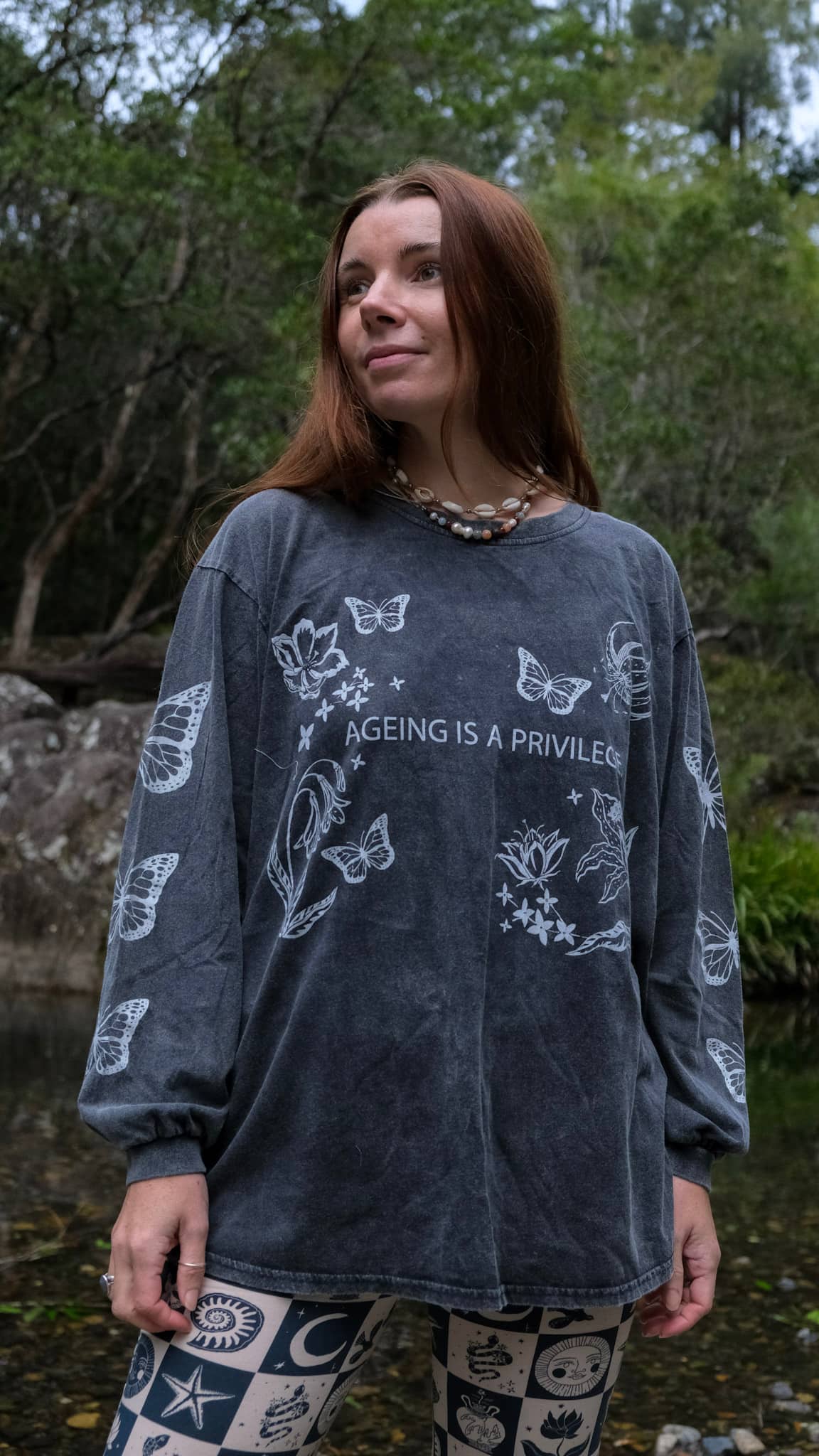 A woman standing outdoors near a stream wearing a dark gray long-sleeve t-shirt with white butterfly illustrations and the text &