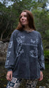 A woman standing outdoors near a stream wearing a dark gray long-sleeve t-shirt with white butterfly illustrations and the text &
