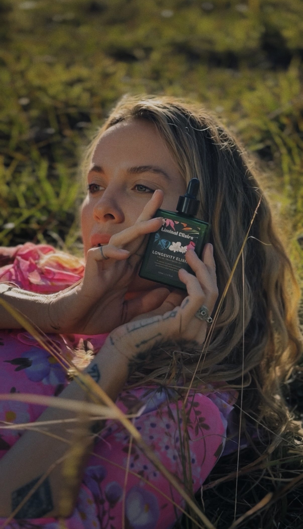 A woman lying in tall grass outdoors holds a dark green bottle of 'Longevity Elixir' skincare product near her face. She is wearing a pink floral shirt.